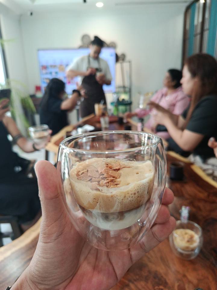 Close-up of a creamy coffee dessert in a glass with blurred café patrons chatting in the background.