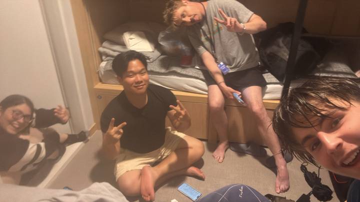 Four hostel roommates sitting on the floor of a compact bunk room flashing peace signs.