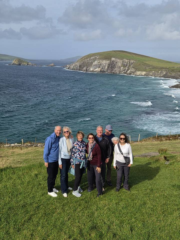Small group of travellers stand on a grassy cliffside above a choppy blue sea and rugged coastline.