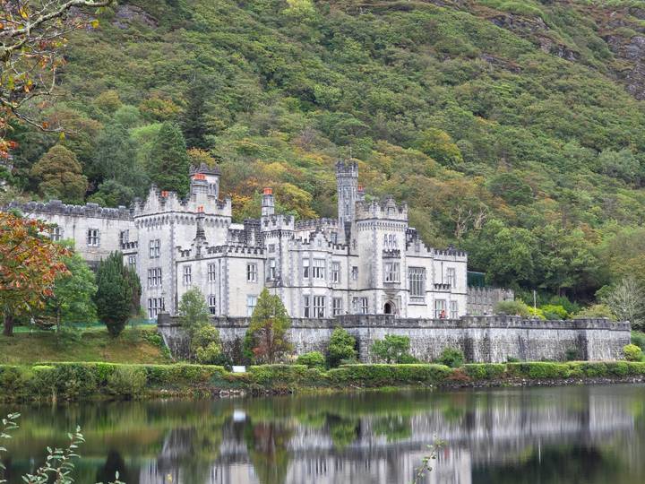 Majestic grey stone Kylemore Abbey reflected in a calm lake backed by forested hillside.