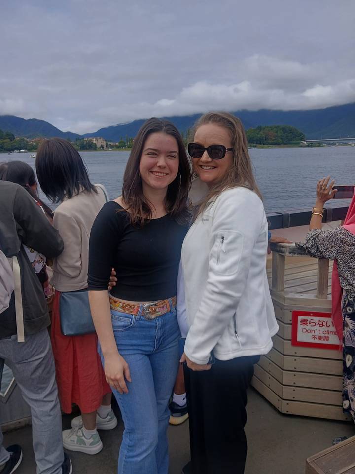 Mother and daughter smile for a photo on a lakeside boat deck with water and shoreline behind.