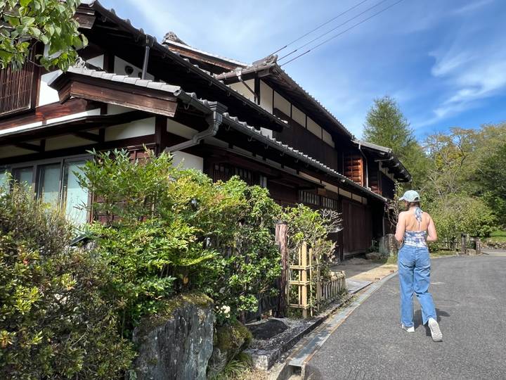 Young traveller strolls past a traditional timber house framed by lush garden plants under blue sky.
