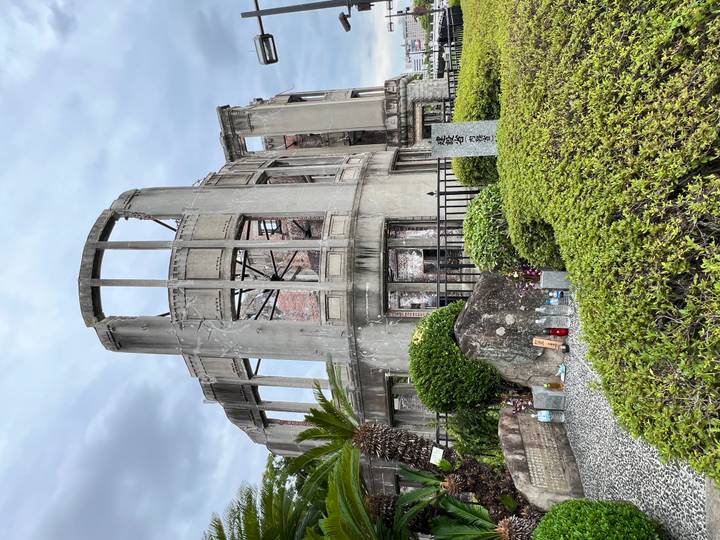 Grey skeletal remains of the Atomic Bomb Dome stand solemnly against a cloudy sky surrounded by greenery.