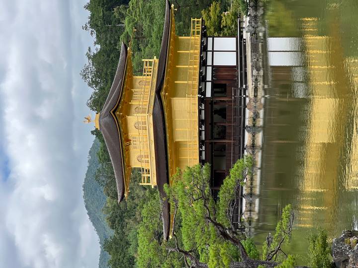 The iconic gold-leaf covered Kinkaku-ji temple reflected in a tranquil pond with forested hills in the background.