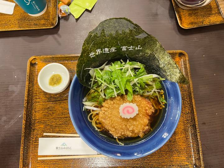 Close-up of a bowl of ramen topped with tempura, fish cake, greens and seaweed on a wooden tray.