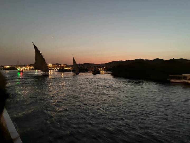 Traditional feluccas sail on a calm river at twilight with city lights reflecting and hills silhouetted.