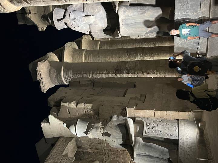 Nighttime scene inside an Egyptian temple courtyard with towering capitals and visitors snapping photos