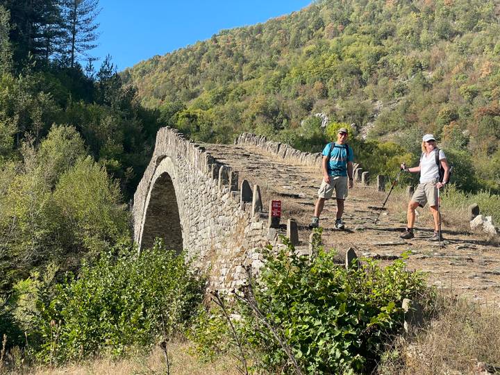 Two hikers posing on top of an old stone bridge surrounded by green hills and autumn foliage