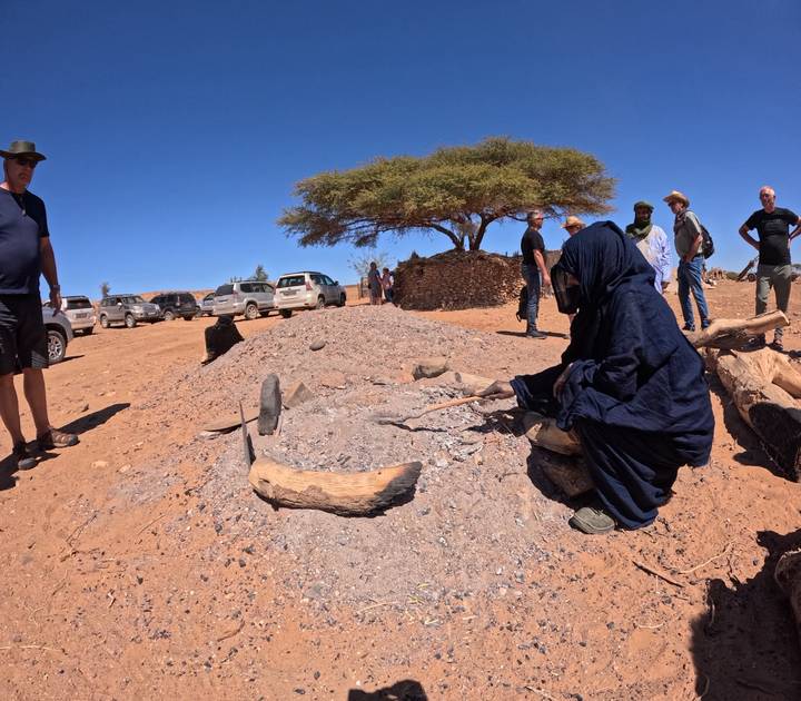 Berber guide in blue robe tending a traditional desert earth oven while travellers watch nearby vehicles