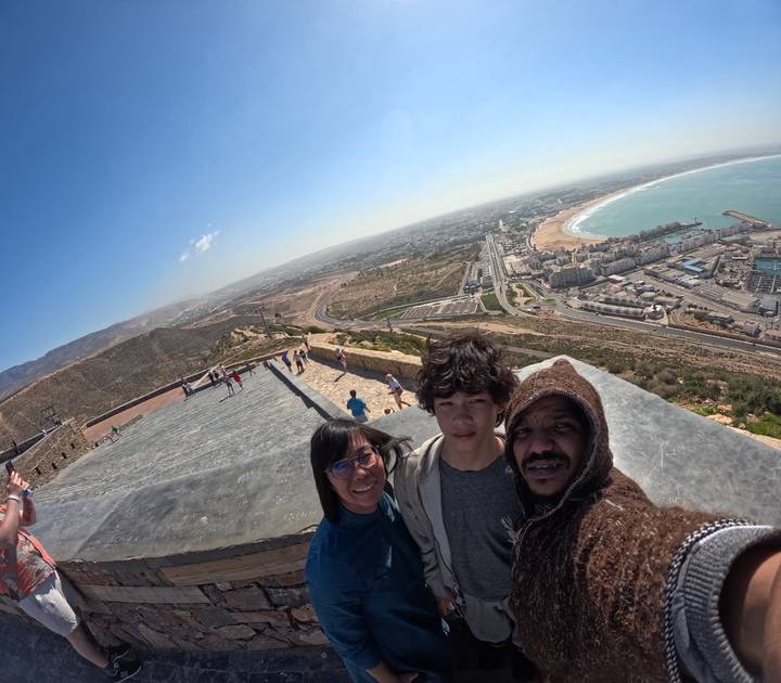 Family selfie on a hilltop viewpoint overlooking a broad coastal city, beach and bay under clear blue skies.