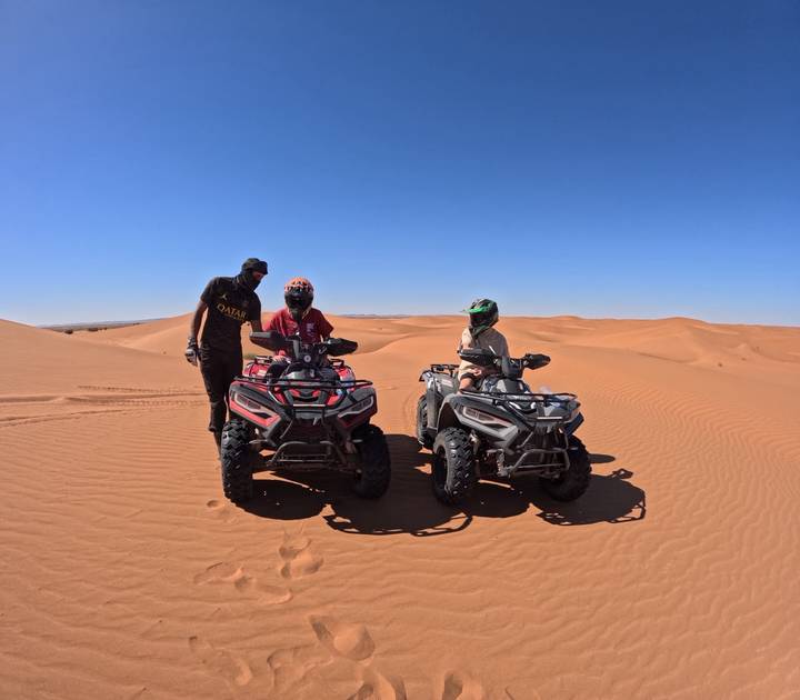 Two quad bikes with riders and a guide stopped on rolling orange sand dunes under a cloudless sky.