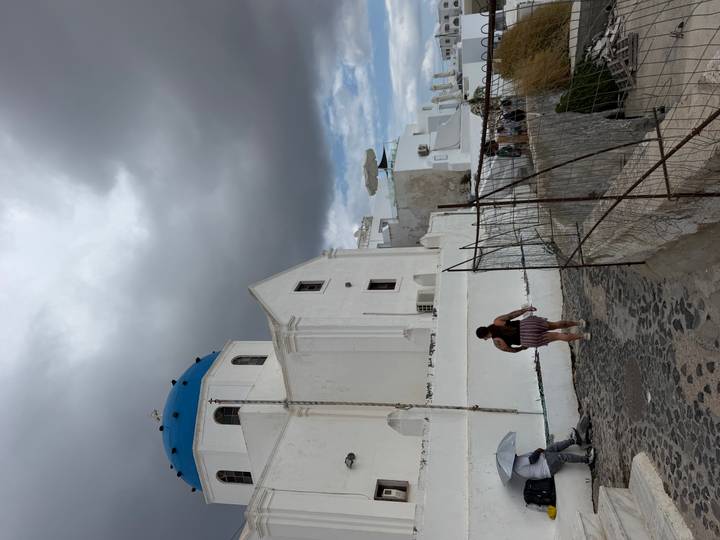 Whitewashed church with blue dome beneath dramatic grey clouds, tourist walking along a cobbled path.