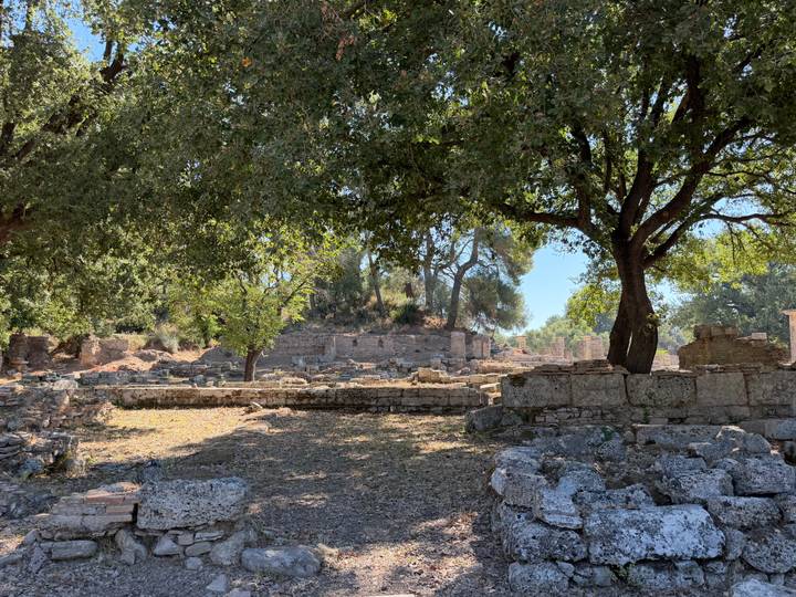 Shaded archaeological ruins framed by large leafy trees with scattered stone blocks.