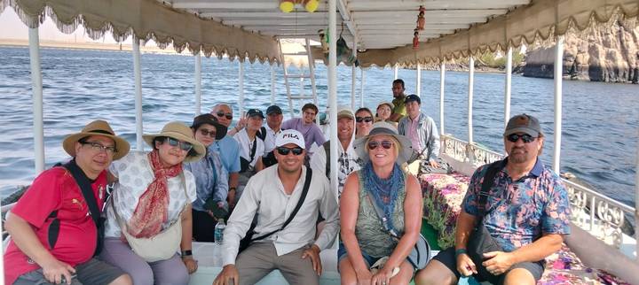 Large tour group sits inside a covered boat cruising on the Nile with blue water visible beyond.