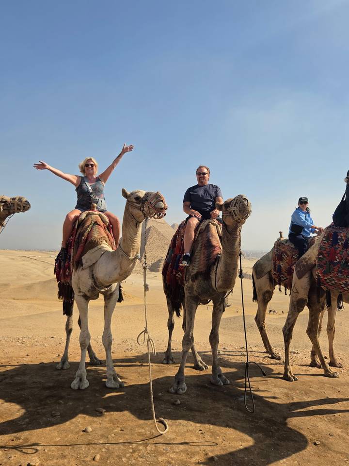 Tourists ride camels in the desert with the Great Pyramid of Giza towering behind under a clear sky.