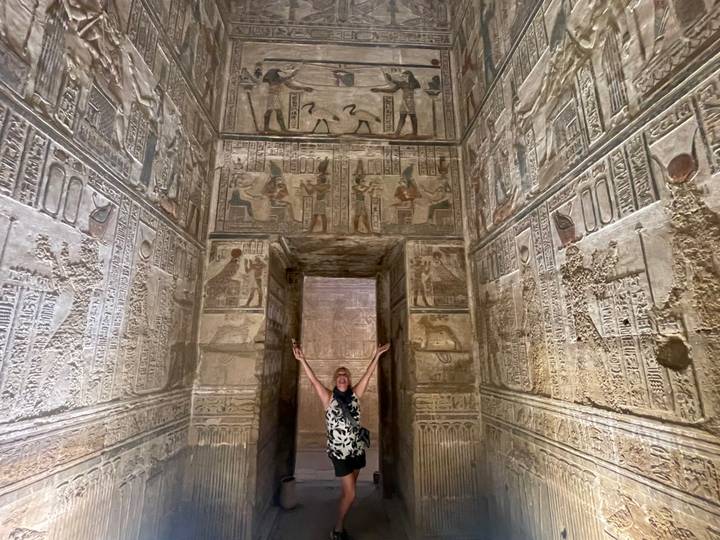 Woman stands with arms raised inside a richly carved and painted chamber of an ancient Egyptian temple.