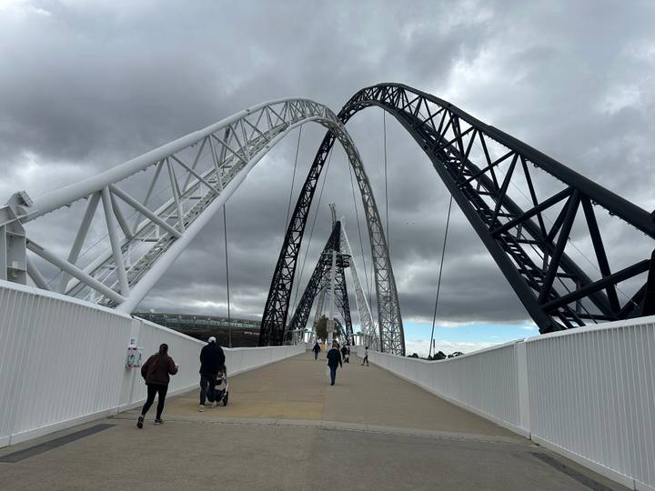 Matagarup Bridge in Perth with dramatic white and black arches spans the Swan River beneath moody clouds and pedestrians.
