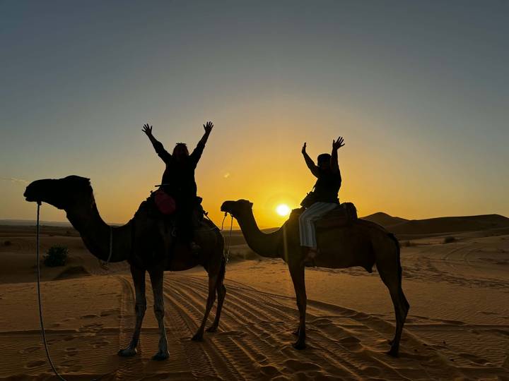 Silhouettes of two riders raising their arms atop camels against a golden Sahara sunset.