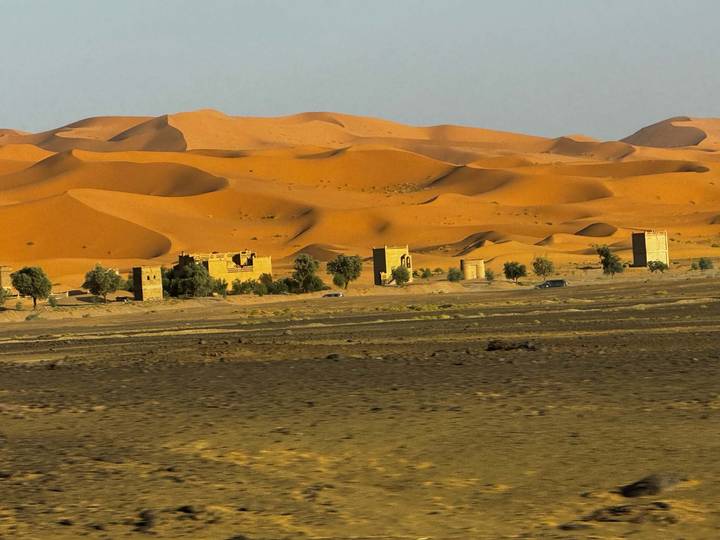 Small desert settlement framed by sweeping orange sand dunes under a hazy sky.