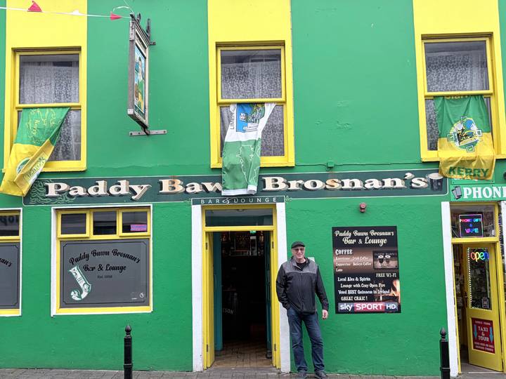 Man stands at the doorway of a vivid green Irish pub façade adorned with county Kerry flags.