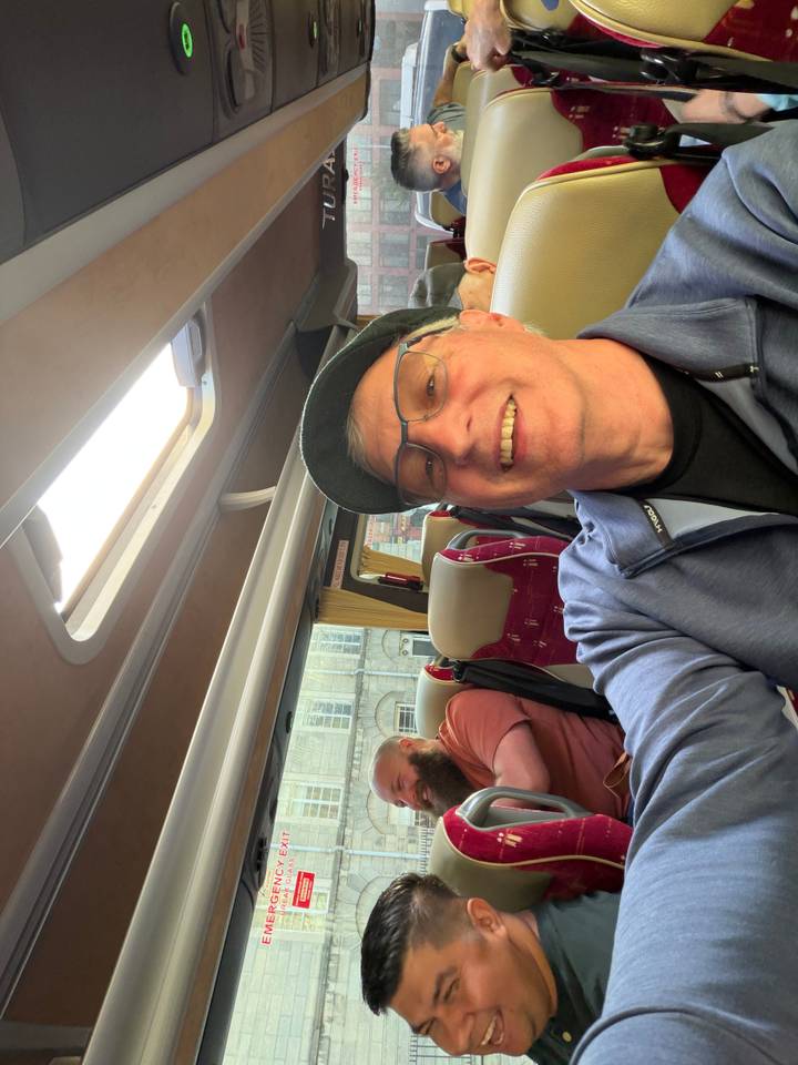 Smiling traveler takes a selfie inside a tour bus with maroon seats and city buildings outside the window.
