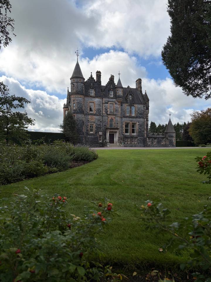 Stately grey-stone manor house with turrets and manicured lawn beneath a partly cloudy sky.
