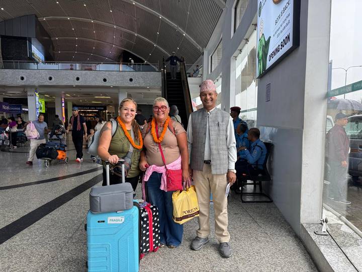 Two female travellers with luggage and marigold garlands stand with a smiling local guide inside an airport arrivals hall.