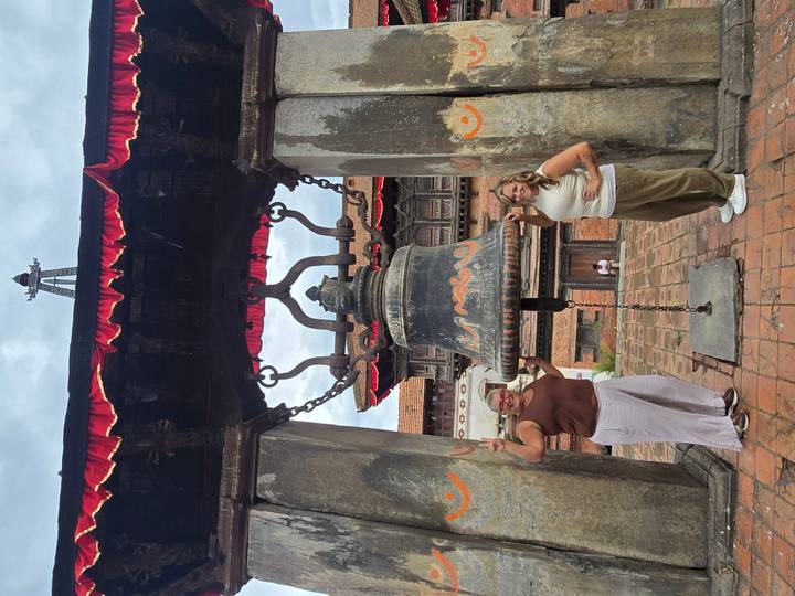 Two women pose on either side of a massive hanging bell in a historic courtyard temple with stone pillars and red trim.