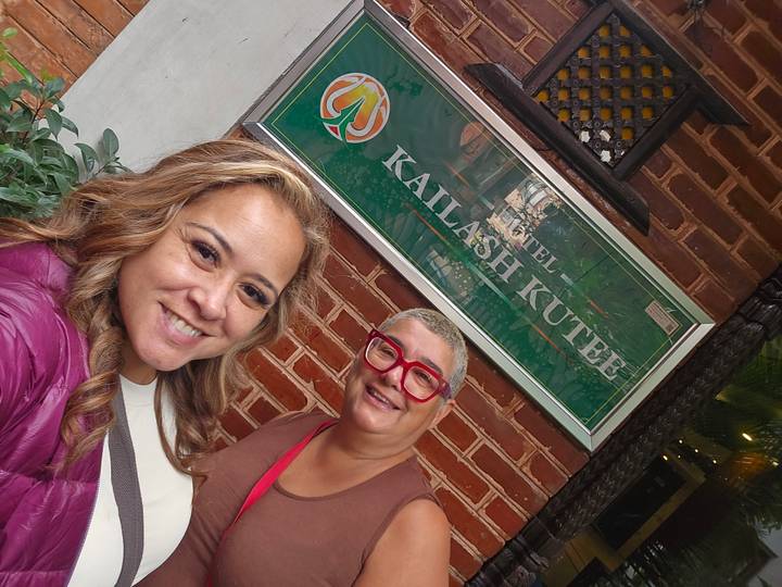Two women take a smiling selfie outside a brick hotel entrance beside a green 'Hotel Kailash Kutee' sign.