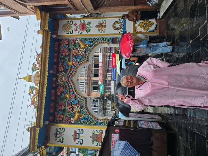 Woman in a pink rain poncho flashes a peace sign beneath an ornate colorful arch while others shelter under umbrellas.