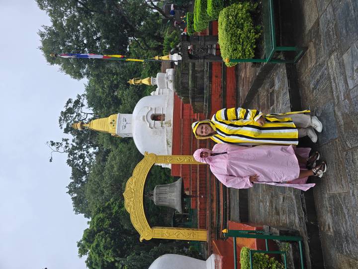 Two travellers in rain ponchos stand before a small white stupa and bell in a green courtyard.