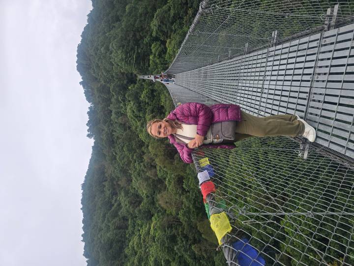 Woman stands on a long metal suspension bridge over a forested valley, prayer flags on the rail.