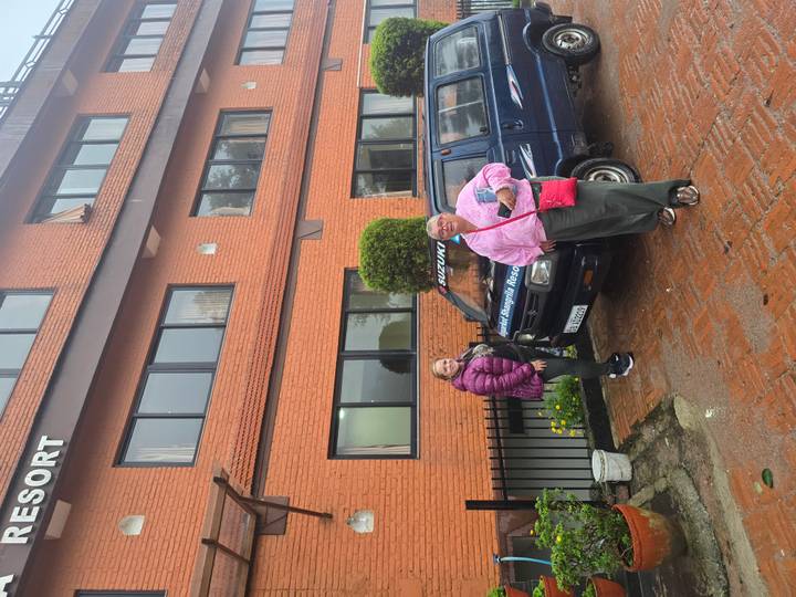 Two women stand beside a blue Suzuki minivan outside a brick resort building on a wet day.
