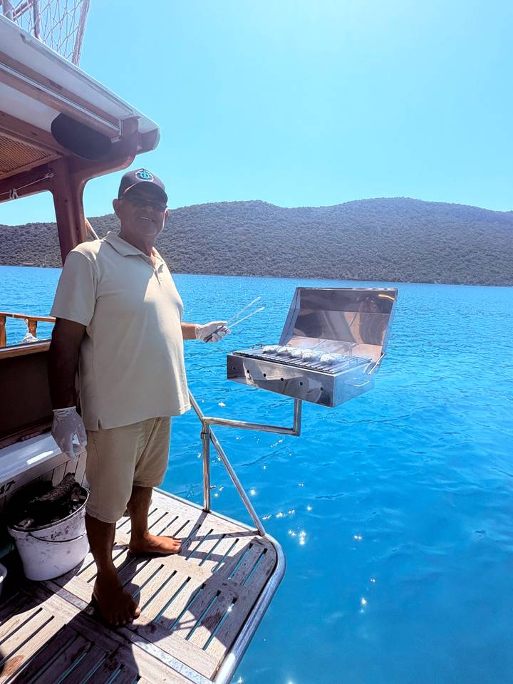 man grilling fish on stainless barbecue at back of boat with bright blue water