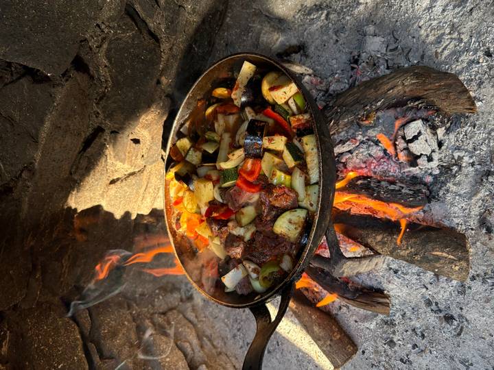 A cast-iron pan full of mixed vegetables and meat simmering over a wood fire beside a stone wall.