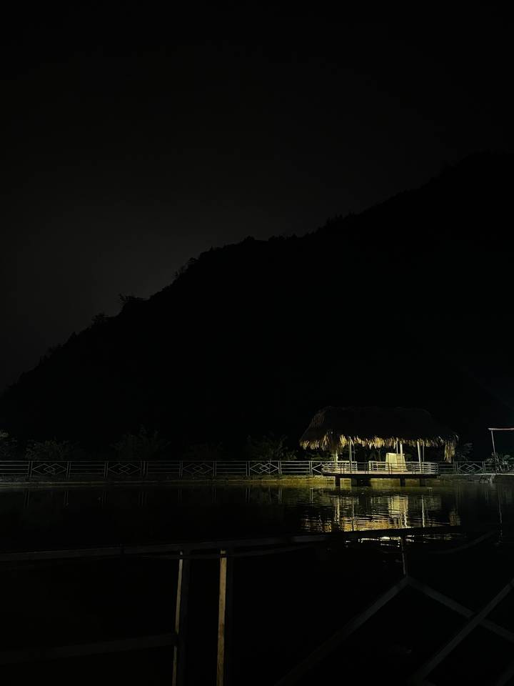 A dark night scene showing the silhouette of a forested mountain and a small thatched shelter lit below.