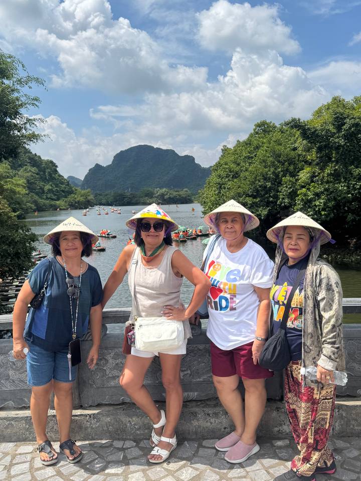 Four women wearing traditional conical hats posing beside a scenic river with karst mountains.