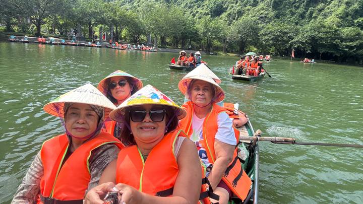 Visitors wearing life jackets and conical hats ride in a wooden rowboat on a green river.