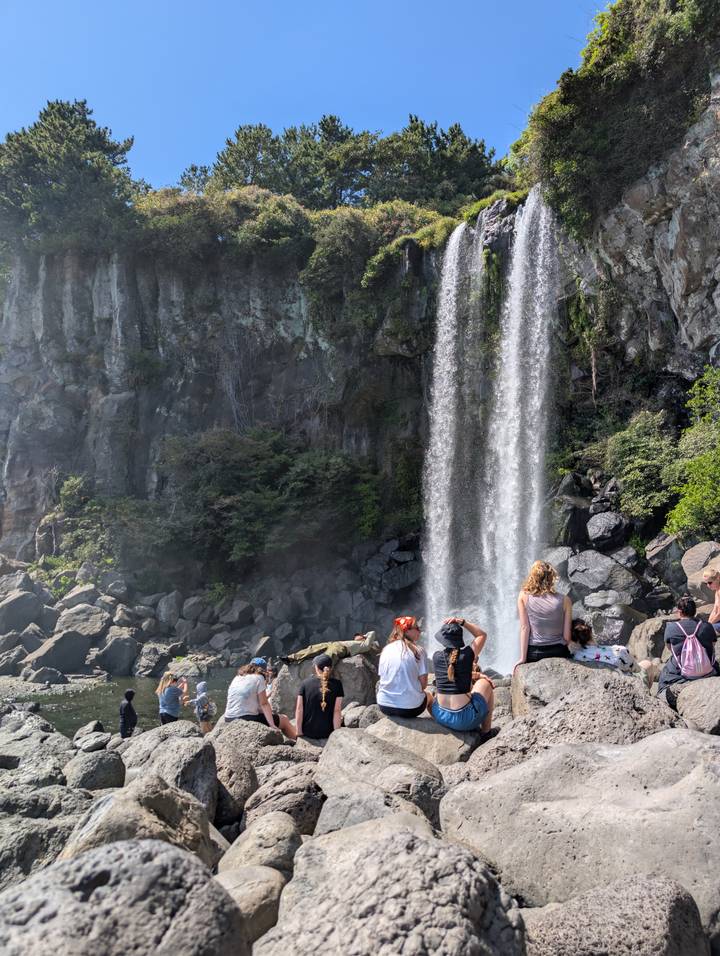Visitors gather at the base of a tall waterfall cascading over basalt cliffs on Jeju Island.