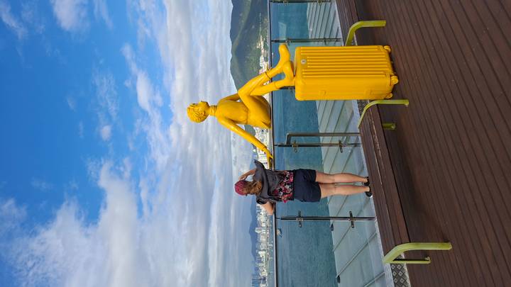 Traveler stands next to a yellow suitcase and bright yellow statue overlooking Busan’s coastline from a viewing deck.