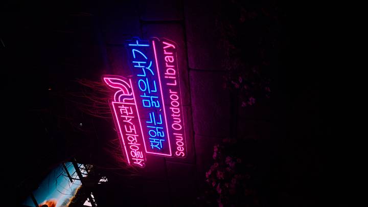 Neon sign in Korean and English reading "Seoul Outdoor Library" glowing pink and blue in the dark.