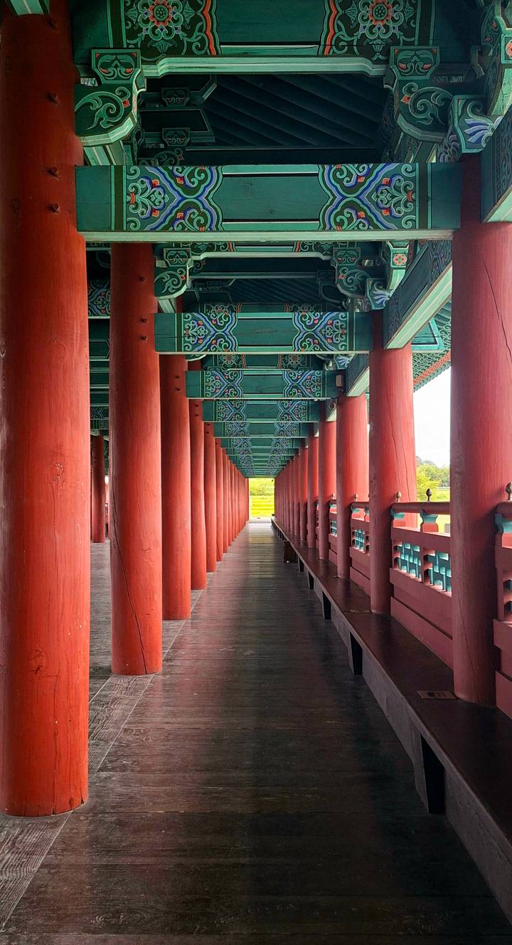 Symmetrical corridor of red wooden columns and ornate green beams in a traditional Korean palace.
