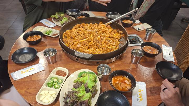 Large pan of spicy stir-fried octopus sits in the center of a table surrounded by diners and Korean side dishes.