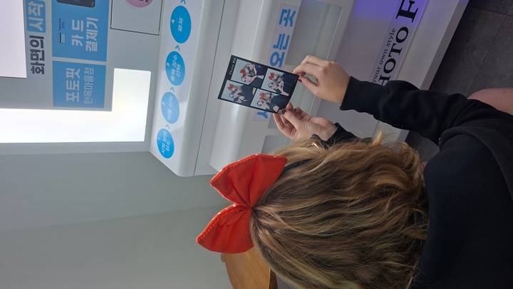 Young woman wearing a red bow headband sits at a photo-booth machine examining a printed photo strip.