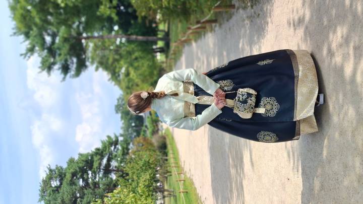 Woman wearing a traditional hanbok strolls down a tree-lined gravel path in a park.