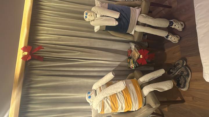 Humorous towel figures dressed as seated passengers with shoes and snacks in a cruise cabin.