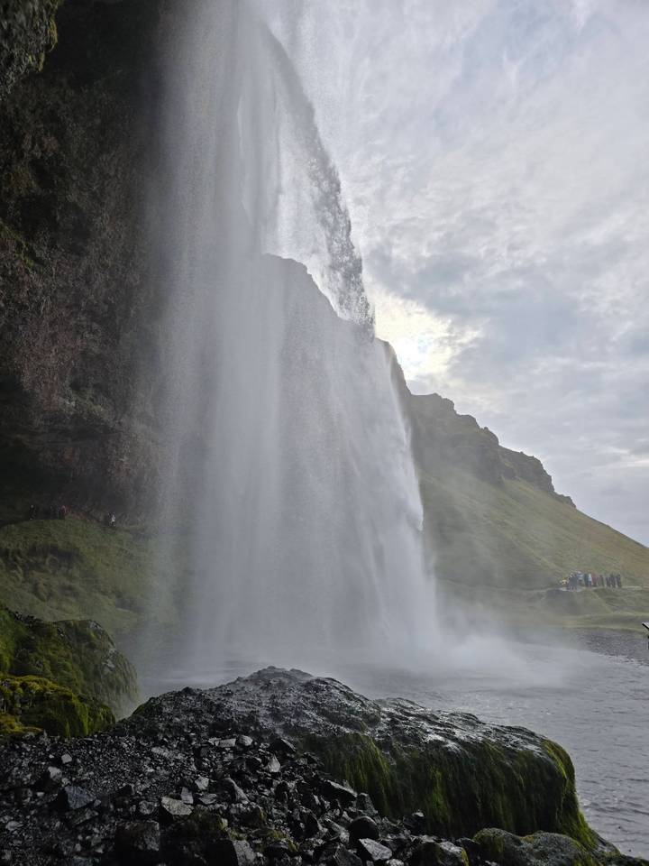 Powerful curtain of Seljalandsfoss waterfall plunging from a mossy cliff with tiny visitors for scale.