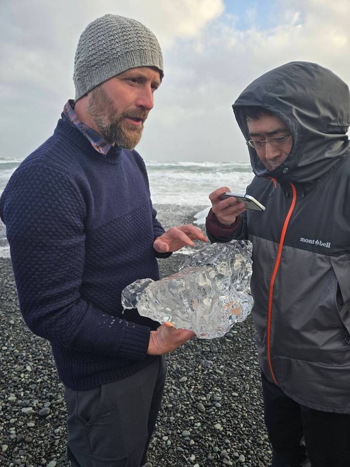 Travellers examine a clear ice chunk on a windswept volcanic beach near crashing surf.