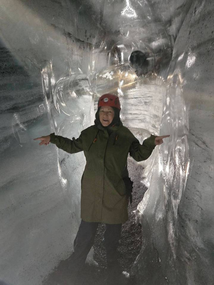 Explorer wearing helmet poses inside a shimmering blue ice tunnel carved through a glacier.