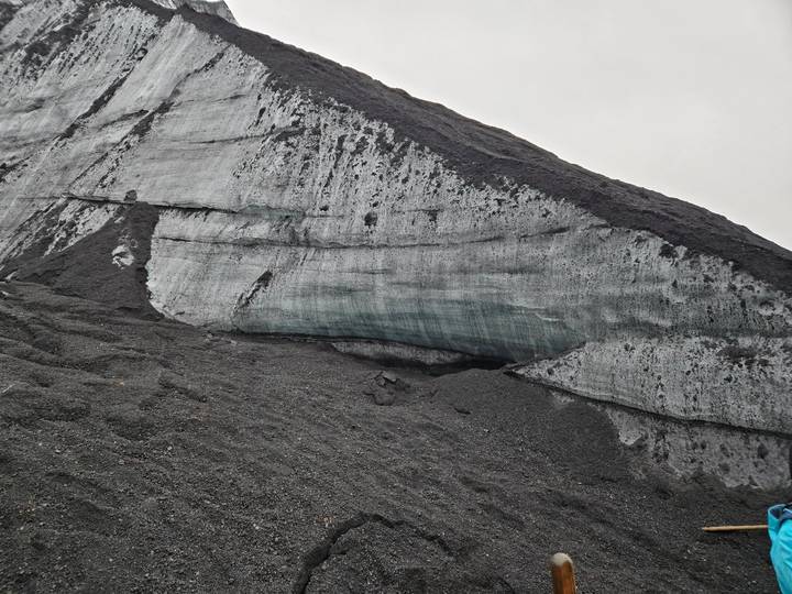 Stark face of a grey glacier with striations and dark volcanic ash layers under an overcast sky.